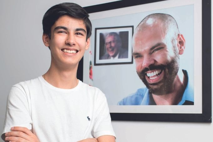 Tomás Covas é um adolescente branco, de cabelos pretos e lisos. Ele usa uma camiseta branca e sorri em frente a um quadro com a foto de seu pai, Bruno Covas, e, mais ao fundo, de seu bisavô, Mário Covas