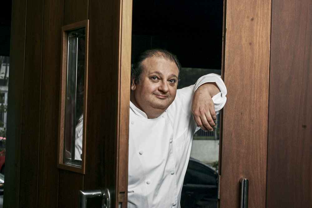 Imagem mostra homem de roupa branca segurando uma porta de madeira, com um dos braços apoiados sobre a parede.