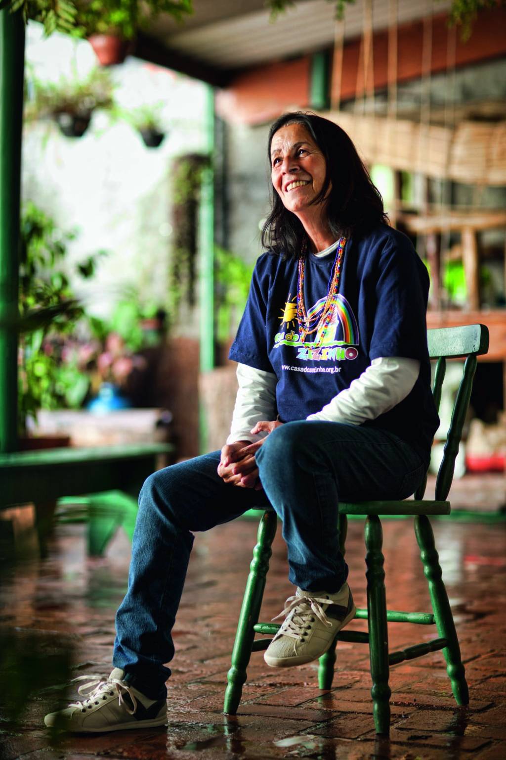 CASA DO ZEZINHO_TIA DAG_DAGMAR RIVIERI GARROUX Imagem mostra mulher sentada em banco verde, vestindo camiseta azul sobre camisa de manga longa branca.