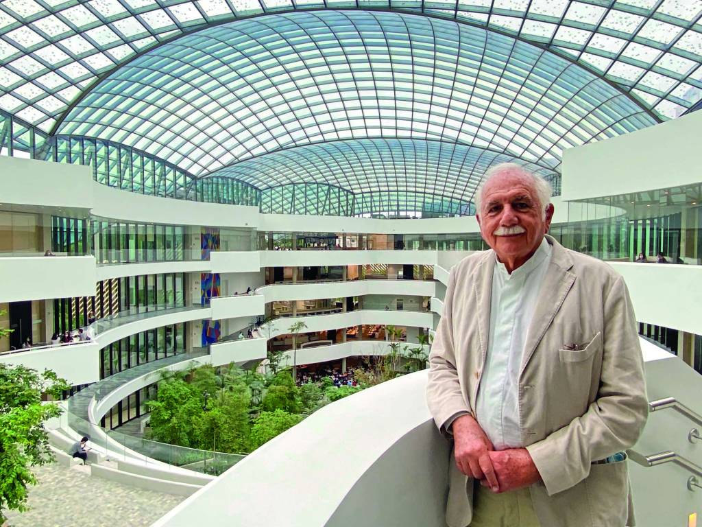 Moshe Moshe é um senhor idoso, de cabelos brancos e bigode branco também. Ele usa um terno bege e posa em uma estrutura onde é possível ver, atrás, a arquitetura do novo prédio do Albert Einstein