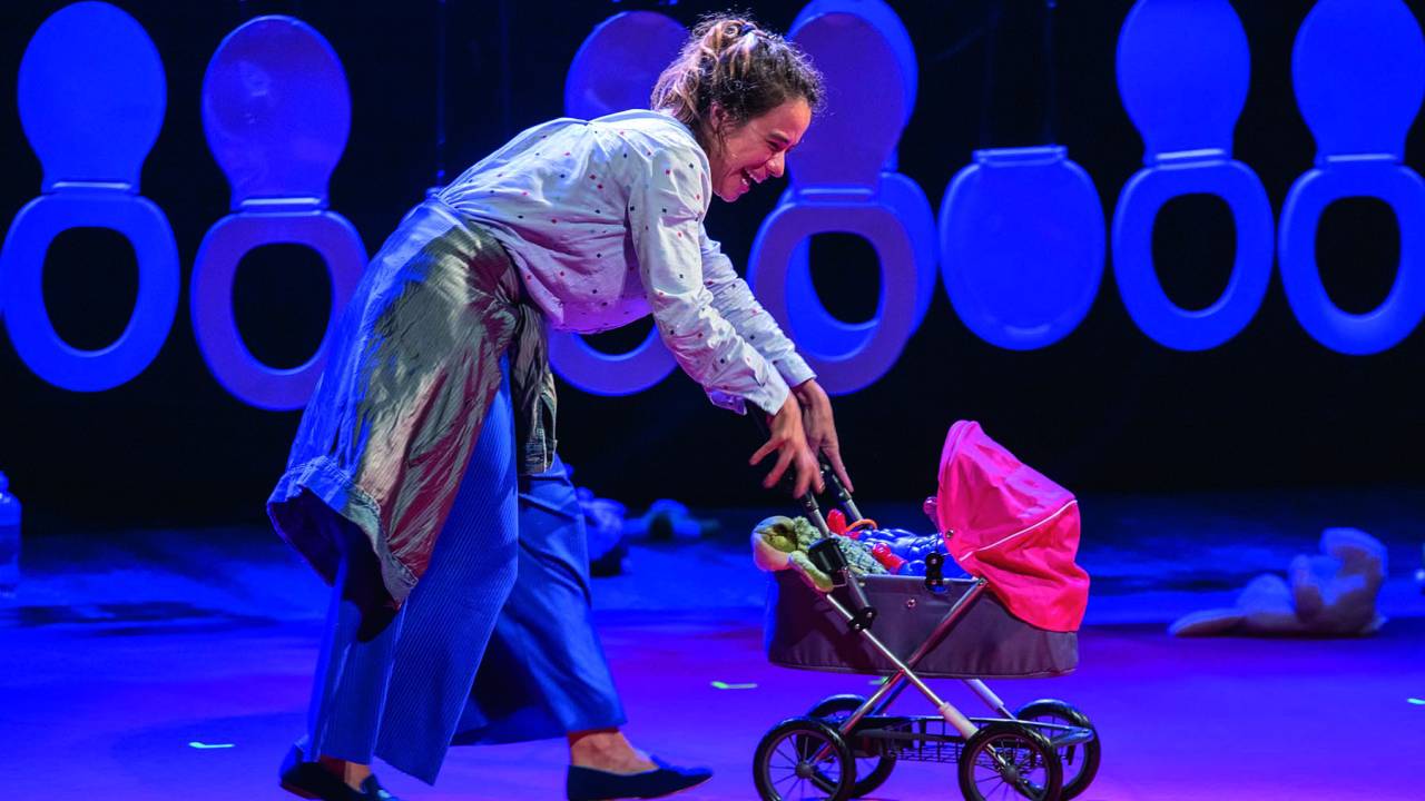 Atriz branca e magra empurra um carrinho de bebê no palco. Ela veste roupas desleixadas e tem o cabelo bagunçado