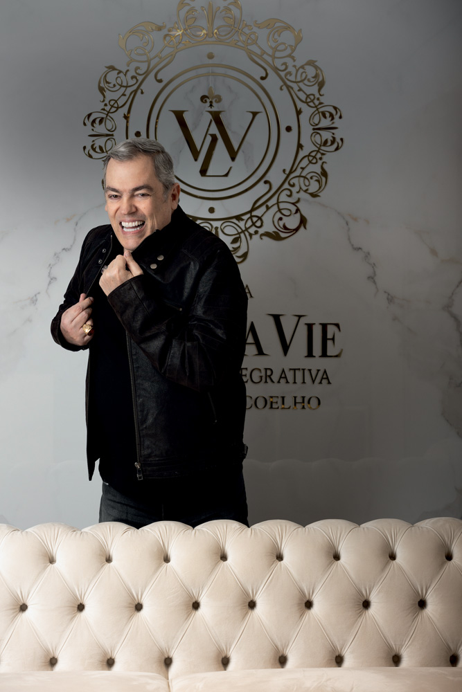 Marco Biaggi Dr. Thulio Coelho Marco Biaggi posa de pé atrás de sofá rosa. Veste tudo preto e segura a gola levantada da camisa.