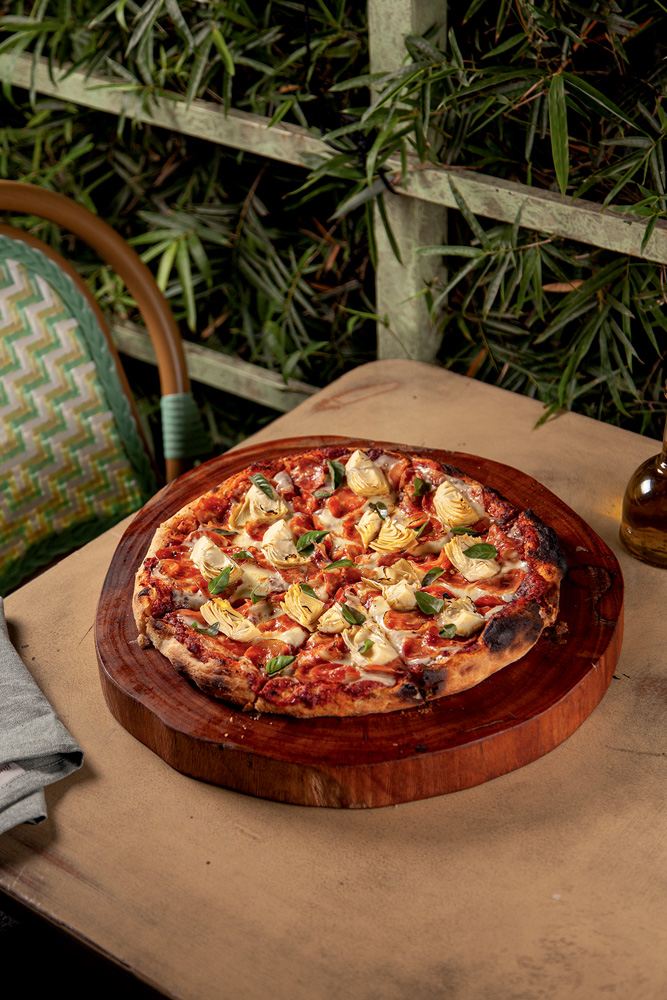 Oli Pães & Pizzas Artesanais Imagem mostra pizza sobre suporte de madeira, em cima de mesa. Ao fundo, folhas e uma cadeira verde.