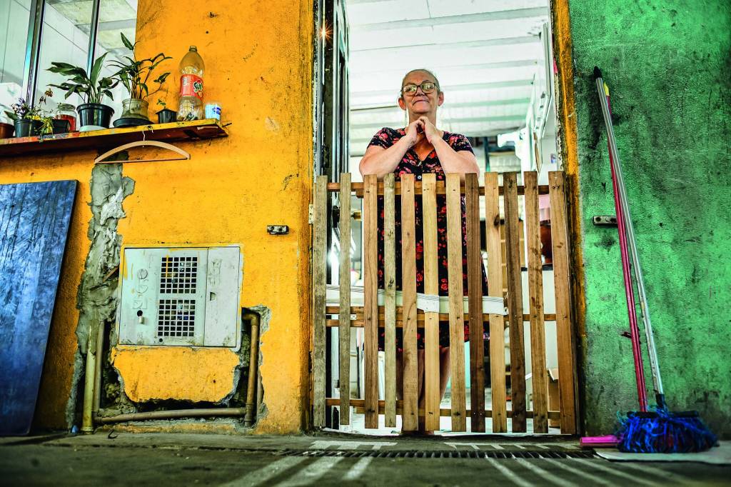 TIAPINGO_FAVELADOCORUJA_4.JPG Imagem mostra senhora atrás de porta de madeira, dentro de casa com paredes amarelas.