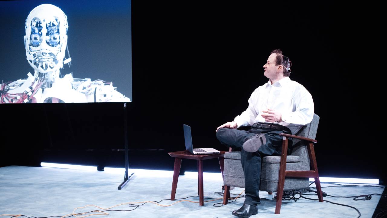 Um robô similar a um homem usa camisa branca, calça jeans e sapatos pretos. Está sentado em uma poltrona no palco, com um notebook em uma mesa a frente. Um telão exibe suas costas, onde ficam expostas as fiações de máquina