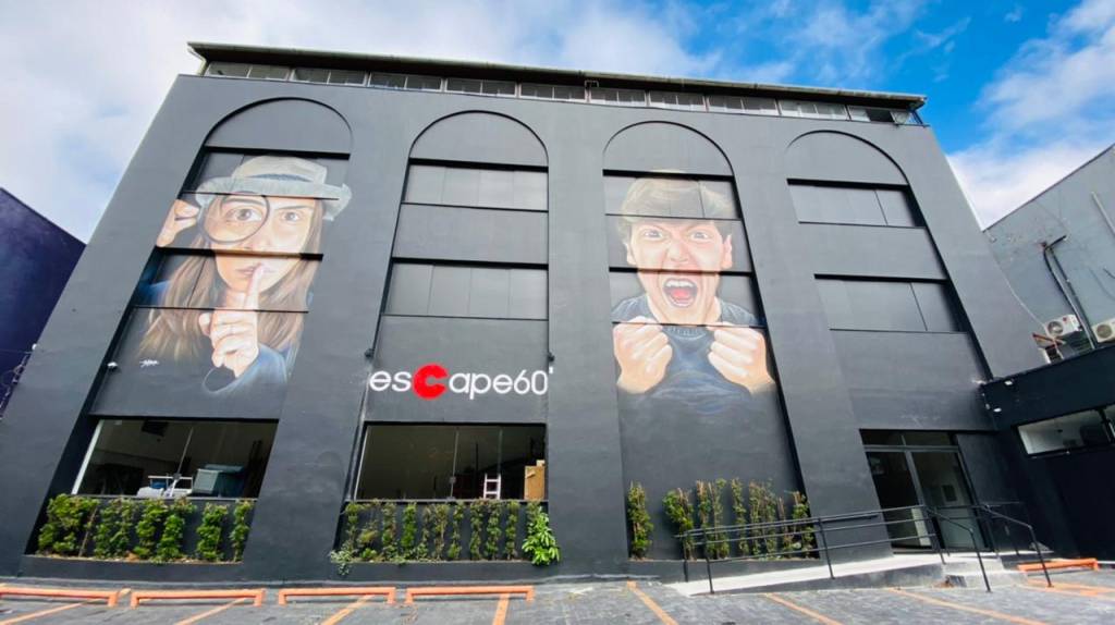 Fachada de uma das unidades da Escape 60, casa de jogos de fuga. É um sobrado preto, com dois detetives, uma mulher e um homem, pintados