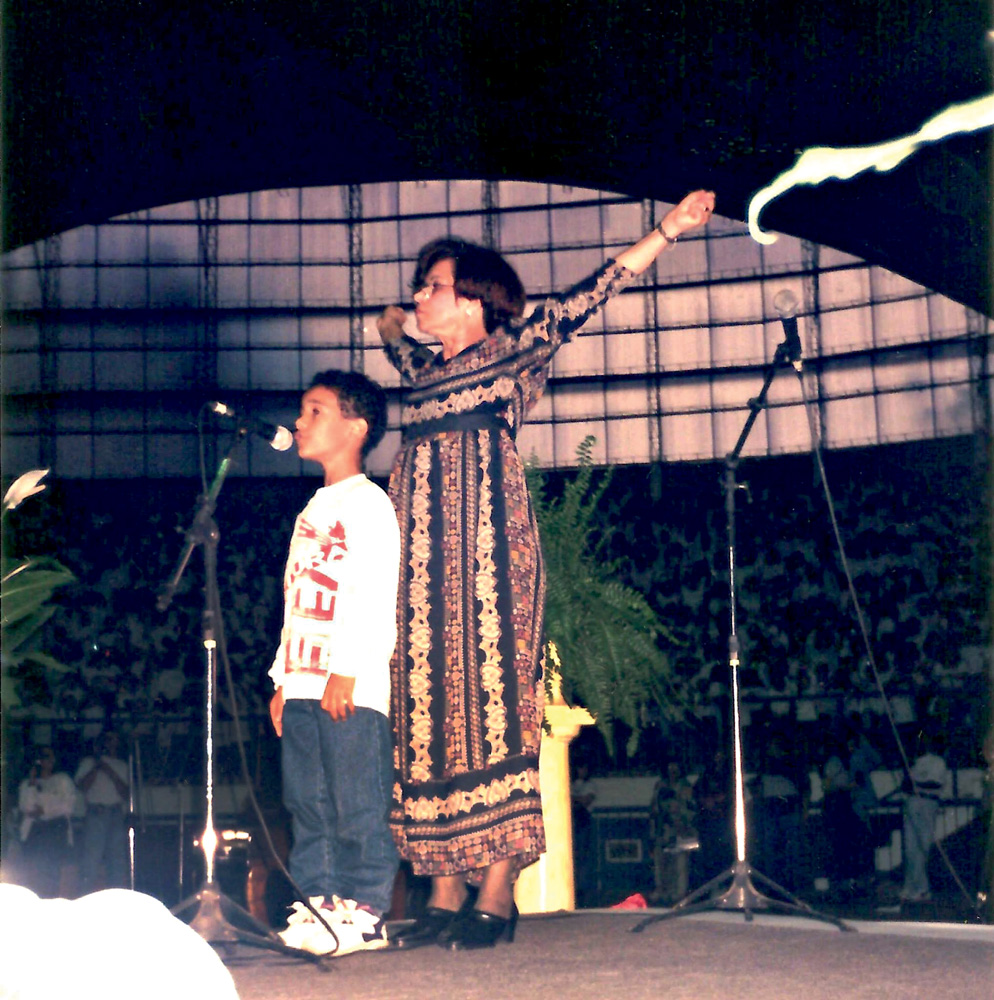 1994_solando com coral de 3000 vozes em Mogi das Cruzes.jpg Imagem mostra criança ao lado de mulher com os braços levantados, em cima de palco, cantando em microfone