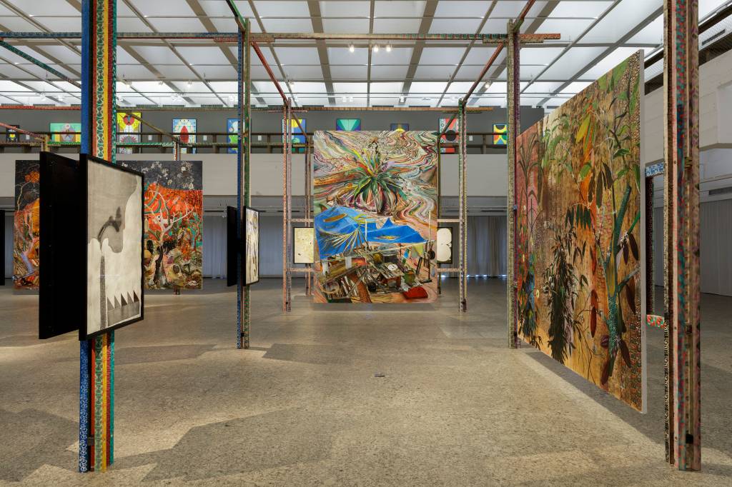 2022_Zerbini_a_mesma_historia_nunca_e_a_mesma_14_Isabella_Matheus Imagem mostra corredor de salão com pinturas coloridas suspensas em estruturas de madeira.