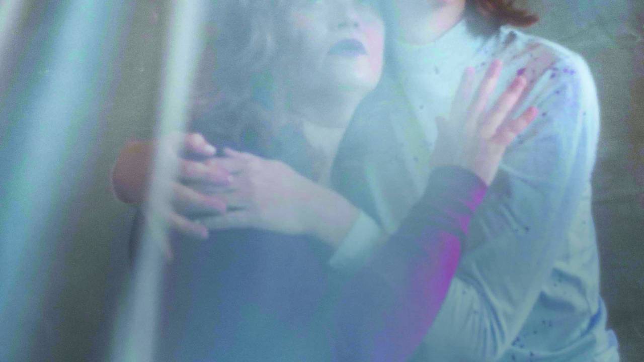 Luciana Schwinden e Bruli Maria: mãe e filha. Duas atrizes brancas, uma ruiva e outra morena, se abraçam envolvidas por um voal