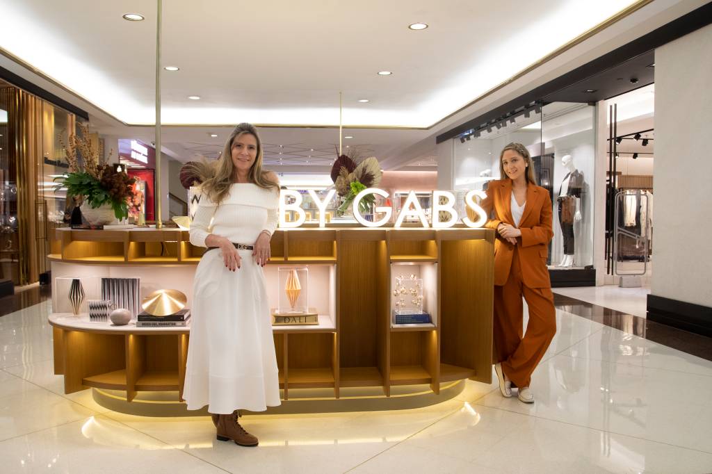 Foto de duas mulheres posando em frente a quiosque de shopping com o letreiro branco "By Gabs"