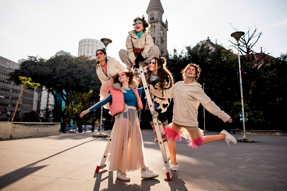 Cinco atrizes, todas brancas, jovens, morenas e magras, vestem figurinos e maquiagens coloridos e se apoiam em escada no Parque Augusta. É um dia ensolarado