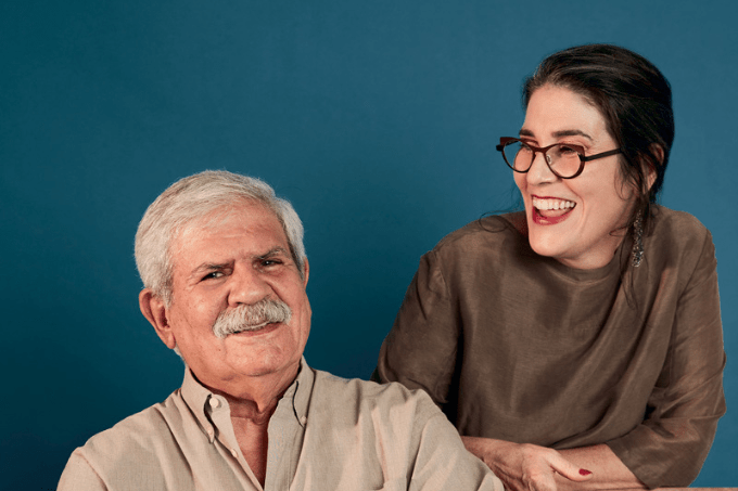 Imagem mostra homem de cabelo e bigode grisalho sorrindo ao lado de mulher de óculos e roupa marrom, sorrindo também