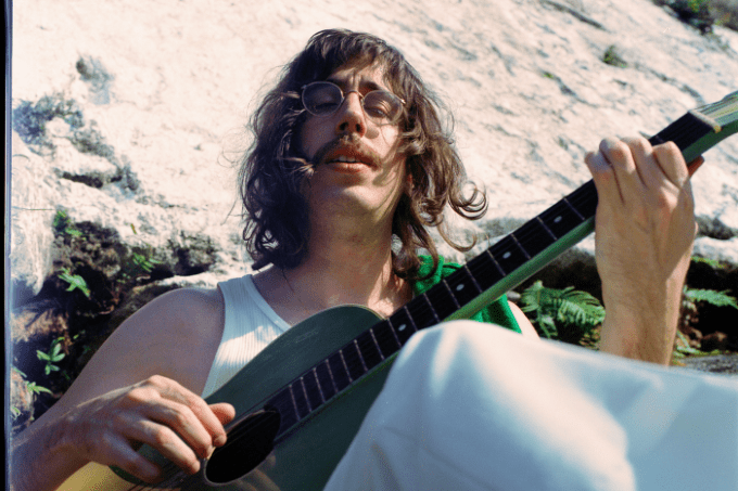 Imagem mostra homem de cabelo comprido e bigode de olhos fechados tocando um violão verde