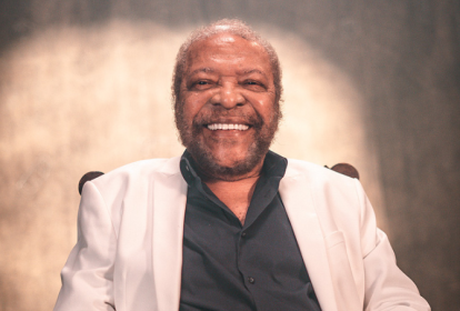 Imagem mostra homem grisalho sorrindo, vestindo camisa preta e blazer branco.