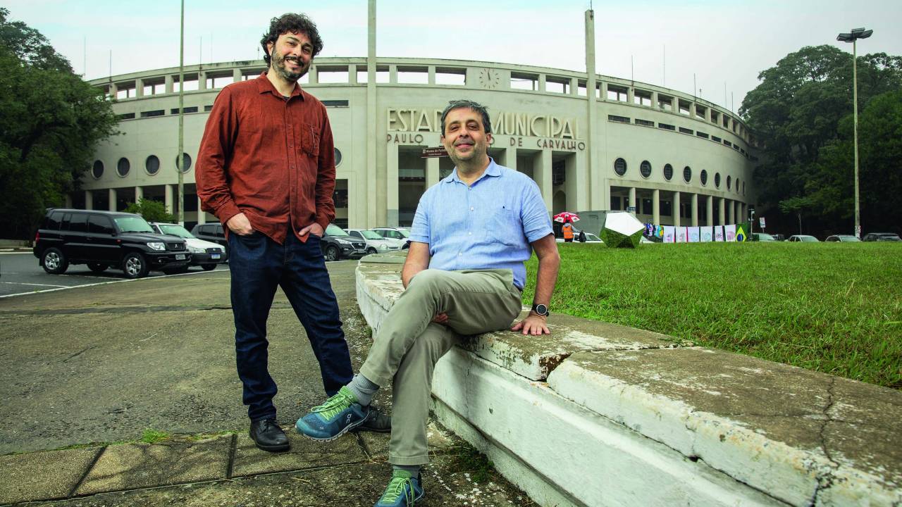 Dois homens brancos e magros, de cabelos escuros em frente ao Pacaembu. O de barba está em pé e o sem barba está sentado em uma estrutura da praça
