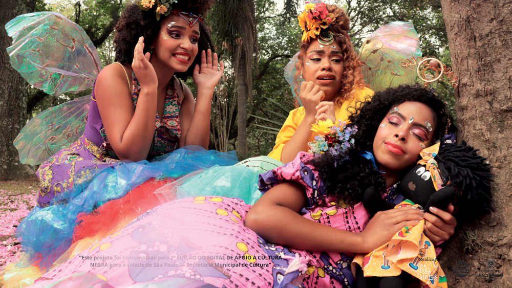 Duas atrizes negras vestem figurino de fadinha e assistem a outra atriz negra, esta vestida de Bela Adormecida, dormir embaixo de uma árvore