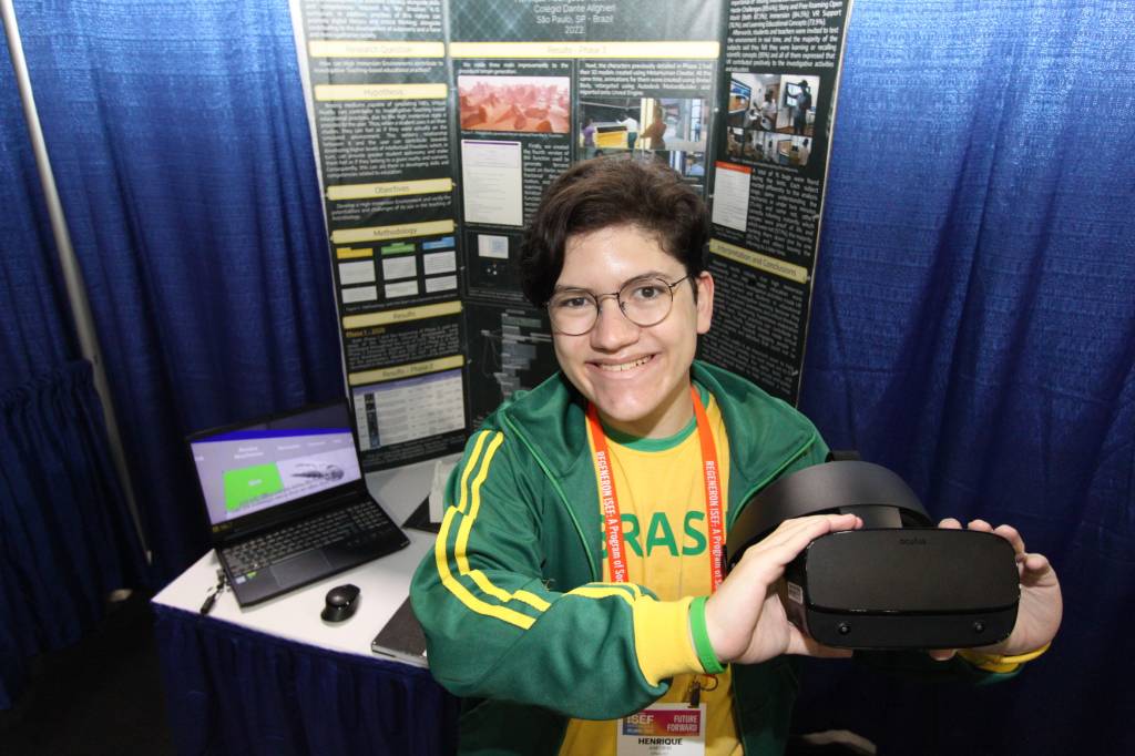 Jovem vestido com jaqueta e camiseta do brasil segura óculos preto de realidade virtual, sorri para a câmera e usa óculos de grau. Atrás dele, um computador notebook.