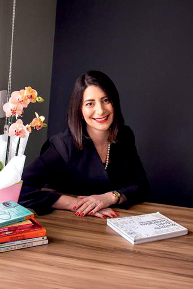 Isabela Nassif.jpeg Isabela Nassif aparece sorrindo sentada a uma mesa e ao seu lado esquerdo há um buquê de flores rosas e laranjas