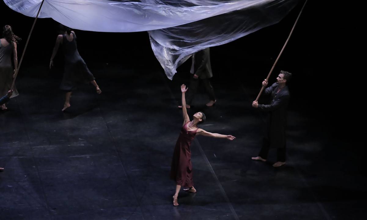 Grupo de dançarinos no palco. Uma dançarina magra, branca e de cabelos curtos e escuros usa um vestido marrom e executa um passo de dança com as pernas cruzadas e os braços para cima, em pé