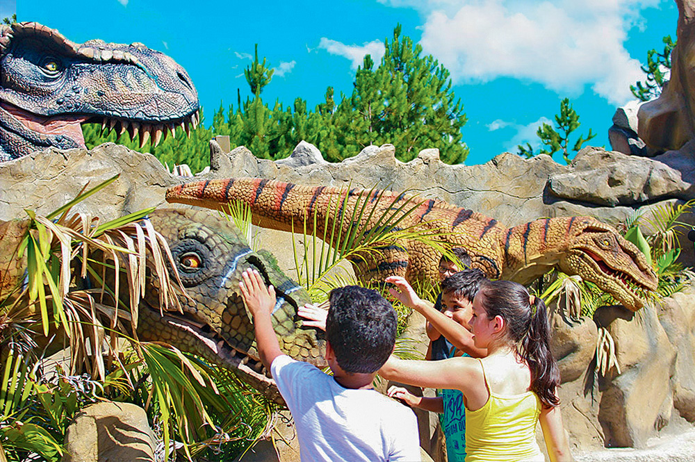 PÁGINA 8 – Acampark (ACAMPARK-DIVULGAÇÃO).jpg Três crianças tocam em uma réplica de dinossauro