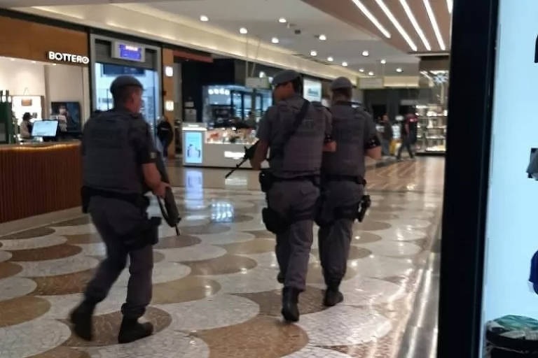 Frame de cena com policiais em corredor de shopping.