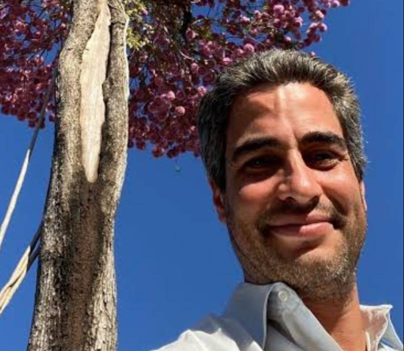 Ricardo Cardim sorri para a câmera olhando de cima. Veste camisa branca e tem cabelos grisalhos. Atrás dele, uma grande árvore com flores rosadas.