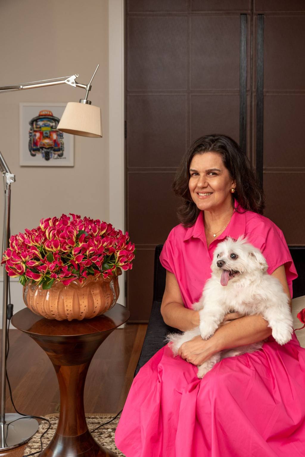 Tetê Castanha (Julia Herman) Tetê Castanha posa sentada com cachorrinho de pelos brancos ao lado de arranjo rosa. Veste vestido rosado também.
