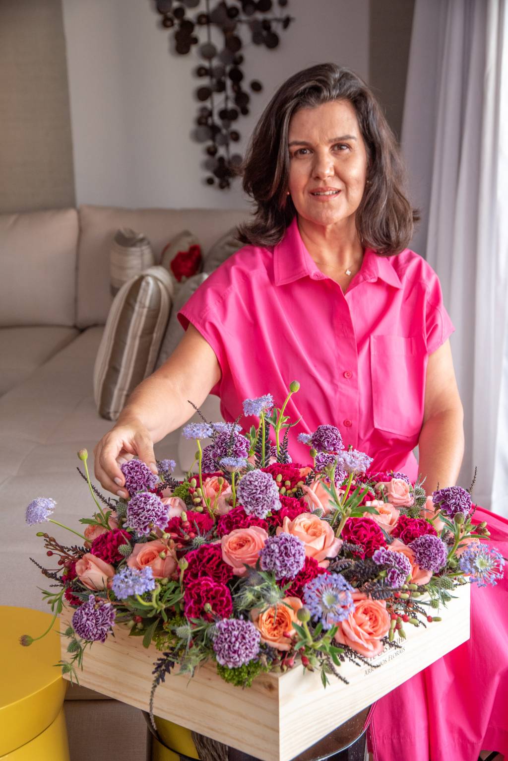 Tetê Castanha (Julia Herman) Tetê Castanha posa vestida de rosa mexendo em arranjo floral com as cores vermelho, roxo e rosa.