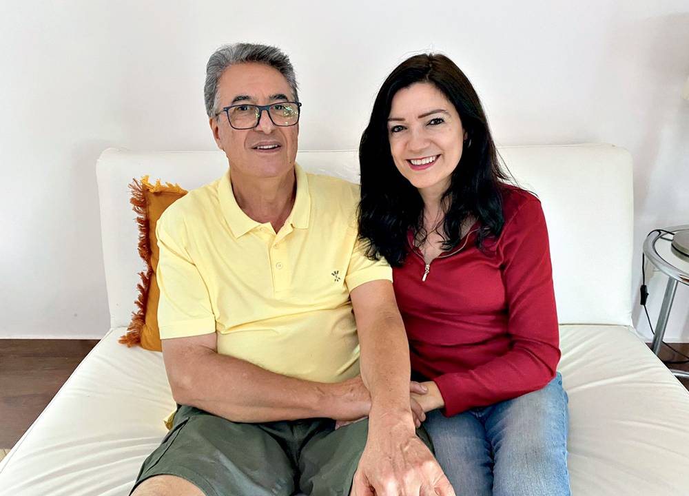 Hélio e Vera sentados um ao lado do outro em uma cama com lençol branco. Ambos são brancos, magros e morenos. Ele usa óculos e é idoso, ela tem cabelos escuros, lisos e longos e é uma mulher de meia-idade