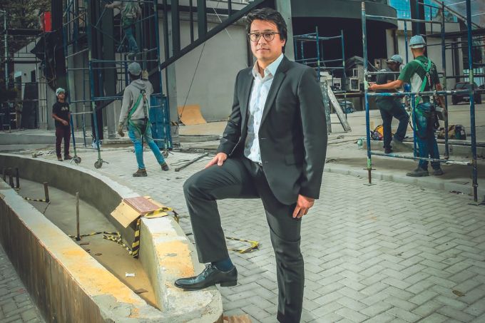 Arena XP Denis Nicolini, 35, idealizador do projeto e um dos cinco sócios da Arena XP, posa com uma perna dobrada em área aberta. Veste terno preto e óculos de grau.