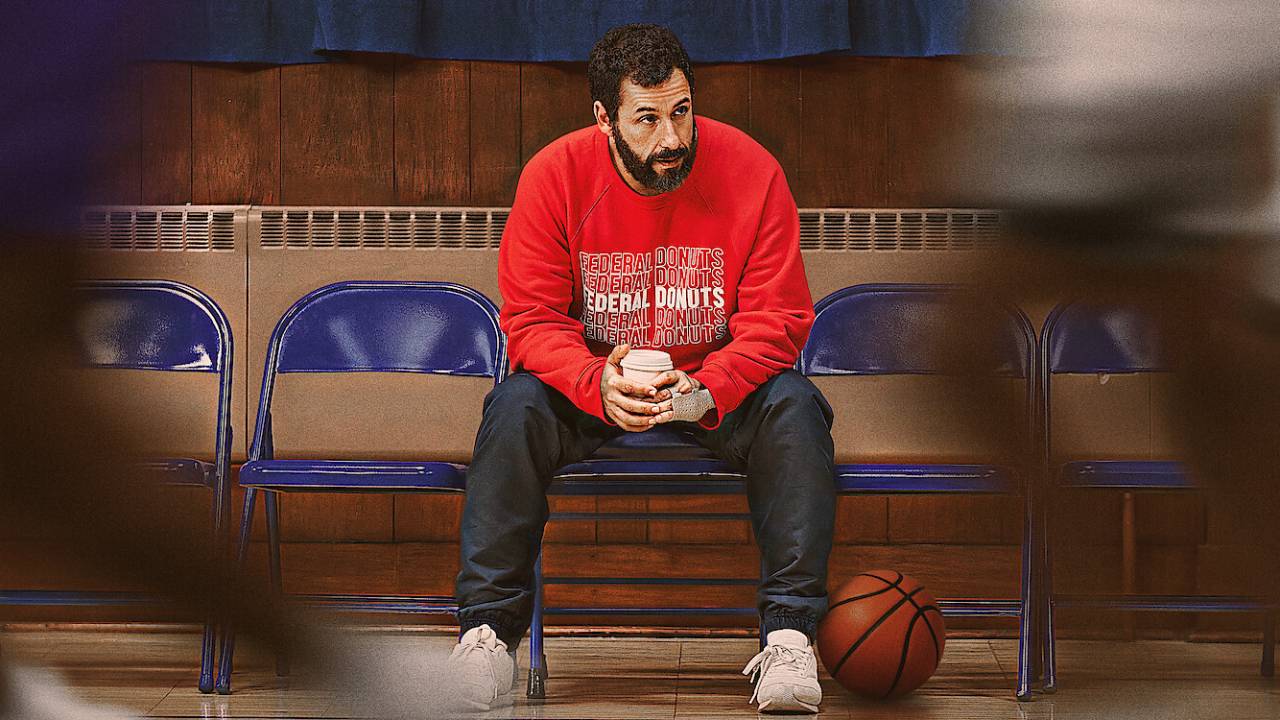 Adam Sandler, de moletom vermelho, observa um jogo de basquete
