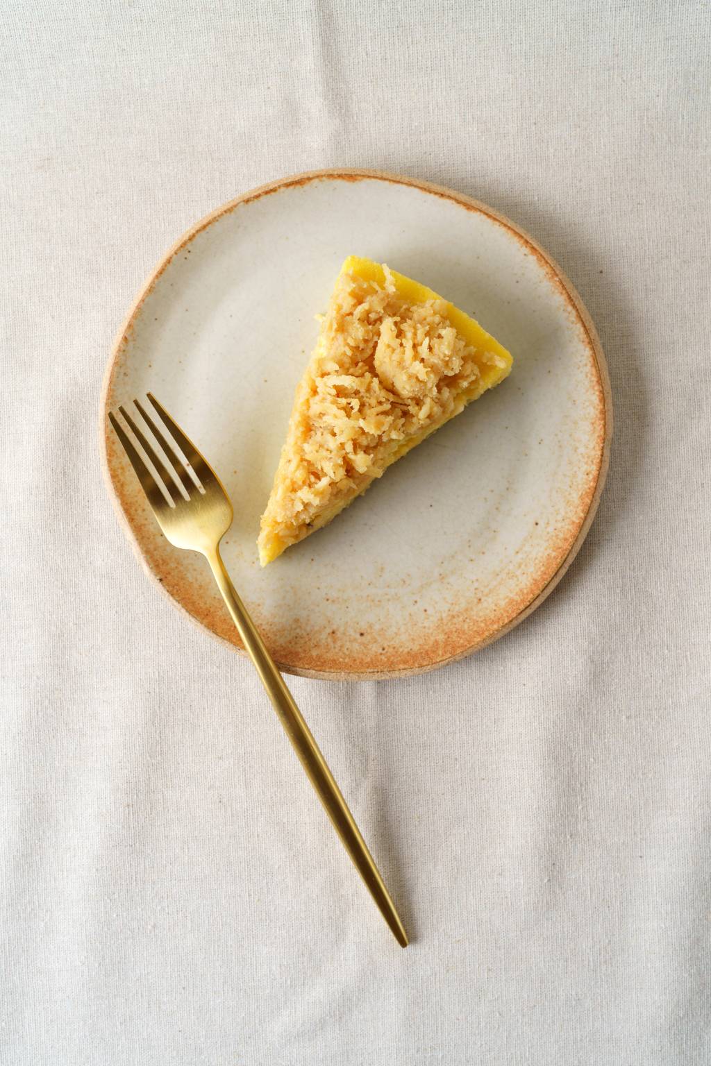Confeitaria Marilia Zylbersztajn Torta de cor amarelada coberta por pedaços de coco queimado servida em prato de cerâmica junto de um longo garfo