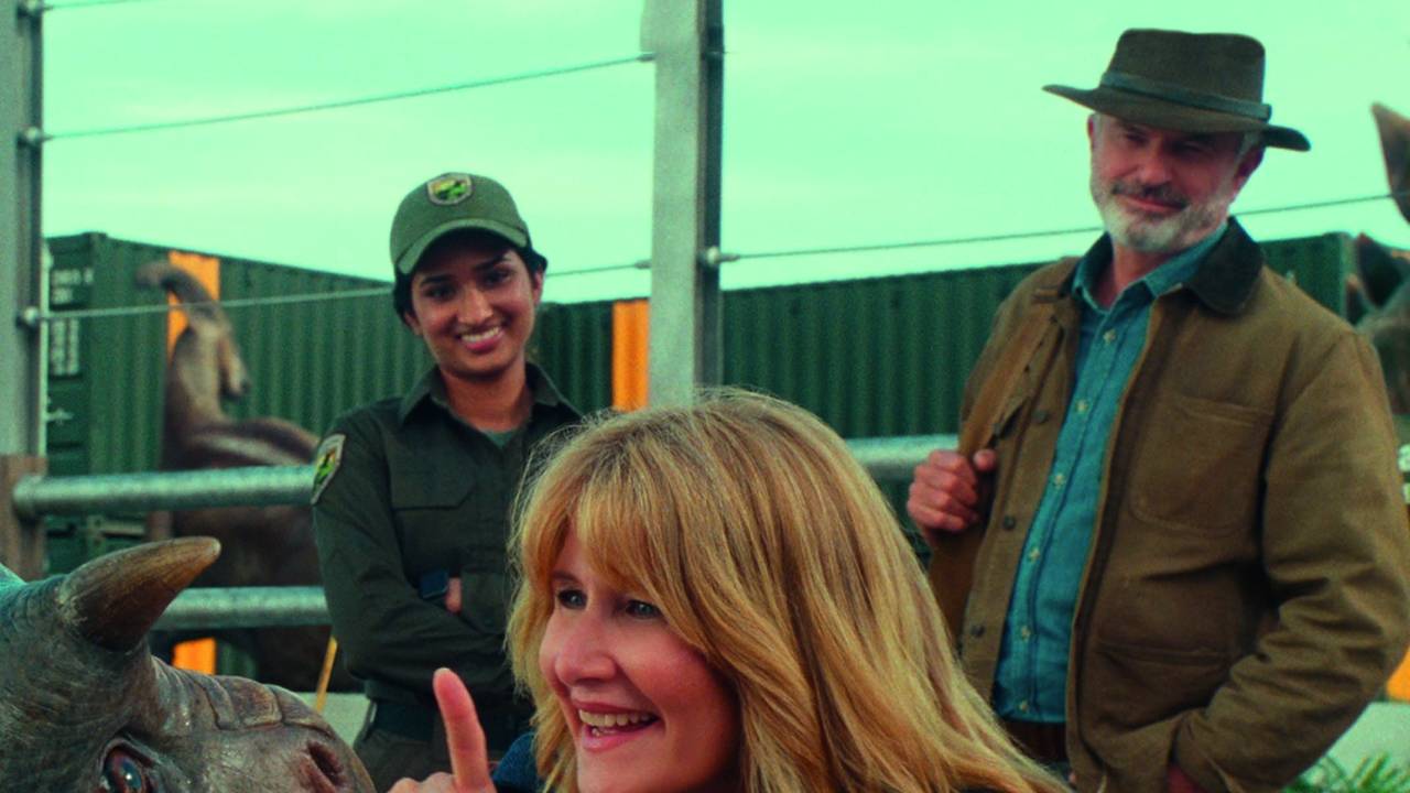 Imagem mostra mulher loira sorrindo, em primeiro plano, agachada em frente a um dinossauro. Uma mulher e um homem, também sorrindo, estão atrás, em pé.