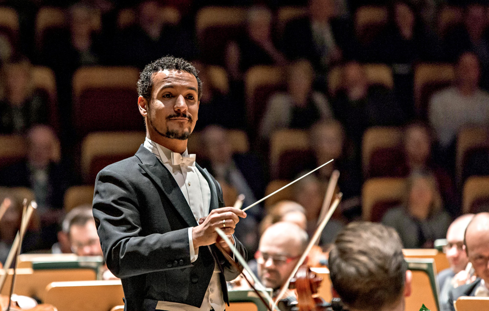 Imagem mostra homem de terno e camisa regendo orquestra