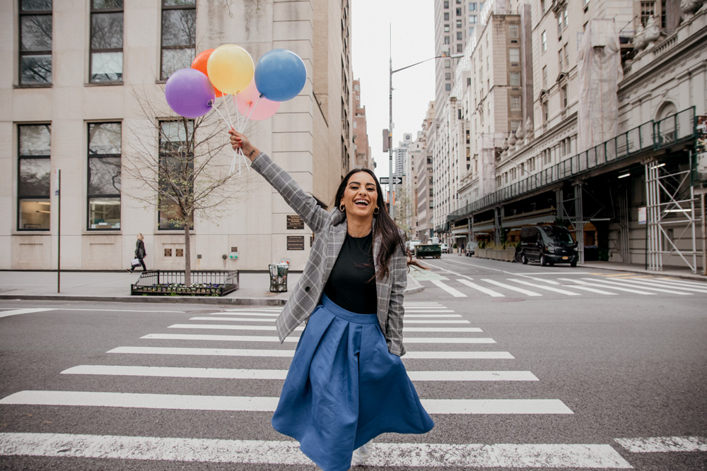Amanda Lima atravessa rua de Nova York sorrindo e segurando bexigas. Veste saia azul, cardigã cinza e camiseta preta.