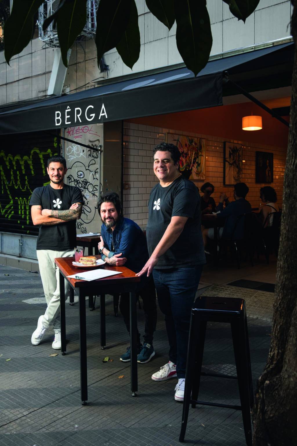 Leo Mereu, Mark Fernandes e Hoffmann, três homens brancos e morenos, de roupas escuras, em frente a um estabelecimento. Um deles está sentado