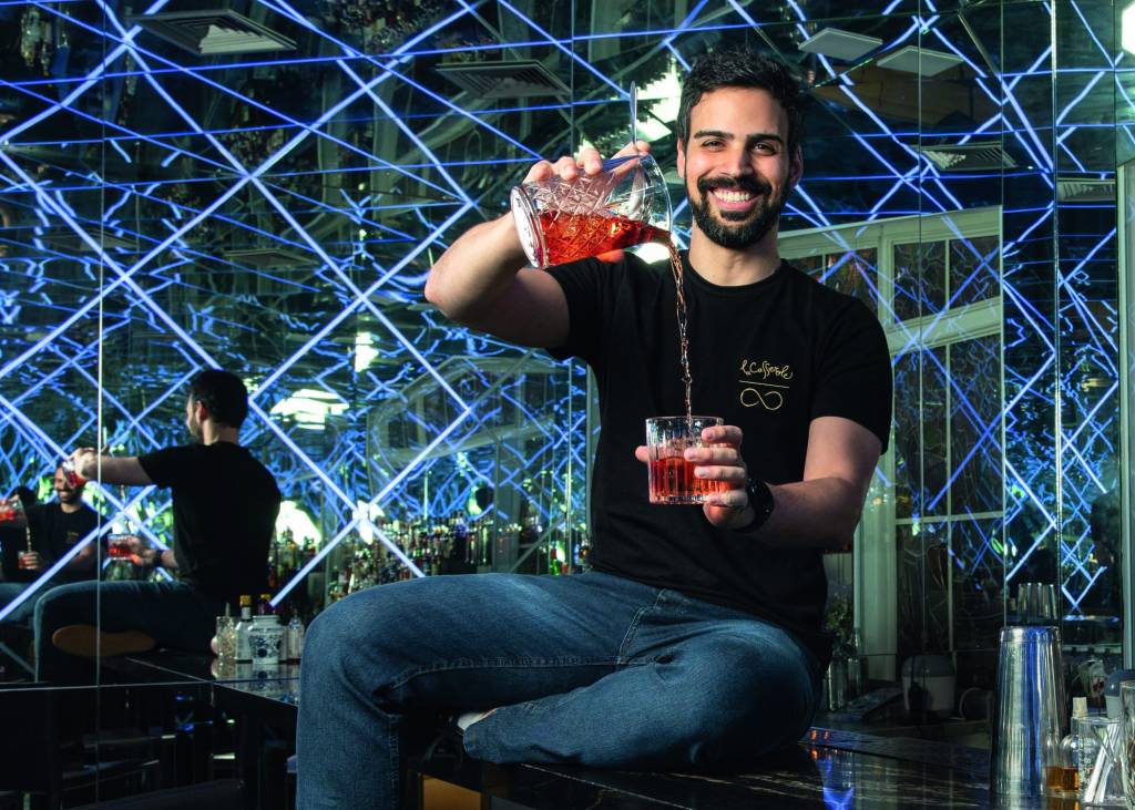 São Paulo, SP 01/07/2022 – Centro SP – Retrato de Leo Henry. Foto: Leo Martins Henry, dono de dois bares, sentado colocando um drinque vermelho de uma jarra de vidro em um copo de vidro. Ele é branco, magro, de cabelos e barba escuros. Sorri para a foto e está sentado dentro de um de seus bares, repleto de luzes azuis