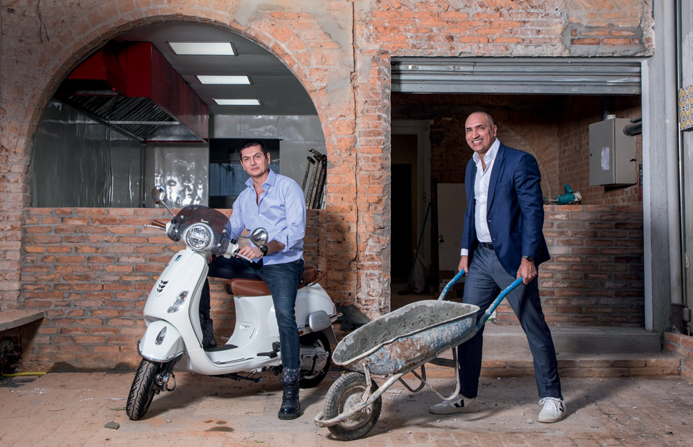 Dois homens, um sentado numa moto e um empurrando um carrinho de construção