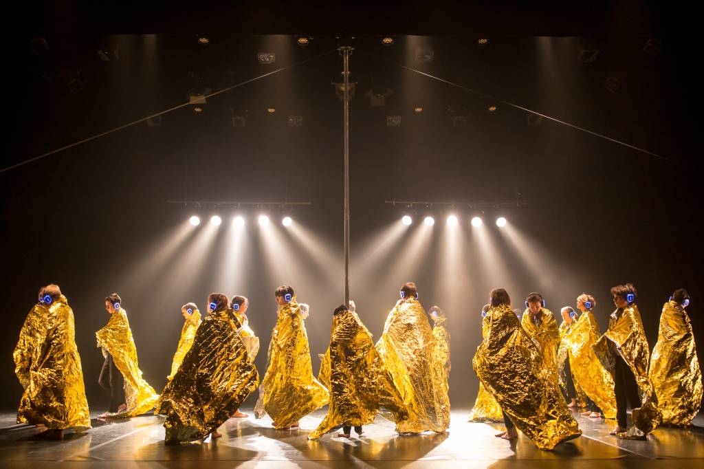 Cie Sam Hester_photo_gregory-batardon Crianças usam capas amarelas e metálicas no palco. São iluminadas por vários feixes de luz branca em cima e usam fones de ouvido tecnológicos
