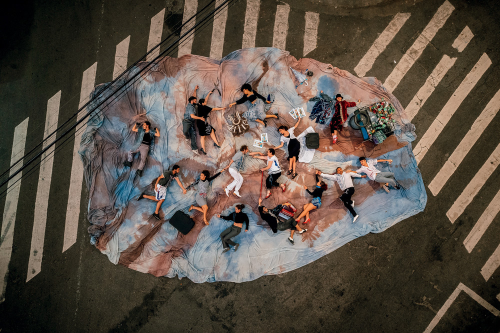 Atores deitados sobre pano azul, com manchas vermelhas. Eles parecem mortos e são vistos de cima, no meio de uma rua vazia