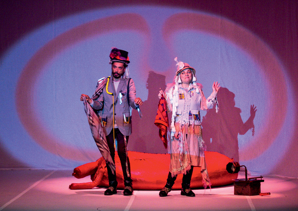 Dois atores com roupas coloridas, feitas de retalhos, em cenário que simula barriga de baleia