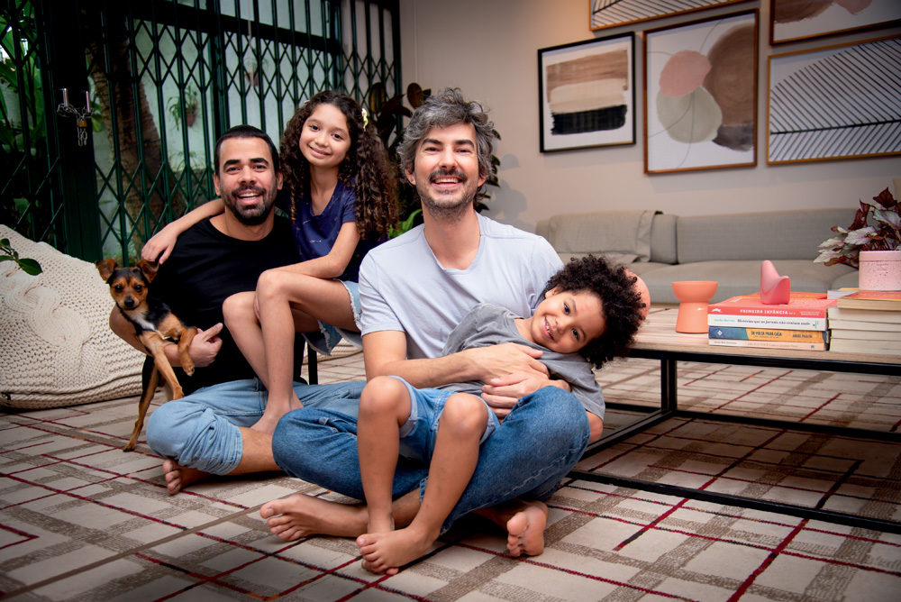 Paulo, um homem branco, magro, de barba e cabelos grisalhos, e Tiago, um homem branco, magro e de barba e cabelos pretos, posam com seus dois filhos sentados na sala de casa. Tiago segura um cachorrinho e Sara, uma menina de longos cabelos cacheados, no colo. Paulo segura Davi, um menino negro, de cabelos crespos e escuros. Todos sorriem para a foto