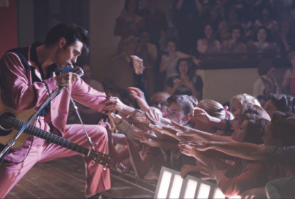 Imagem mostra homem de topete e gel no cabelo em cima de palco, vestindo terno rosa, estendendo a mão para multidão na plateia