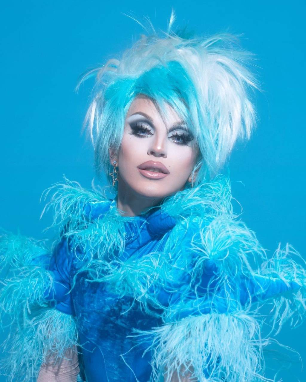 A drag queen Aquaria aparece toda de azul com plumas e cabelo alto. Está com maquiagem bem branca e batom nude.