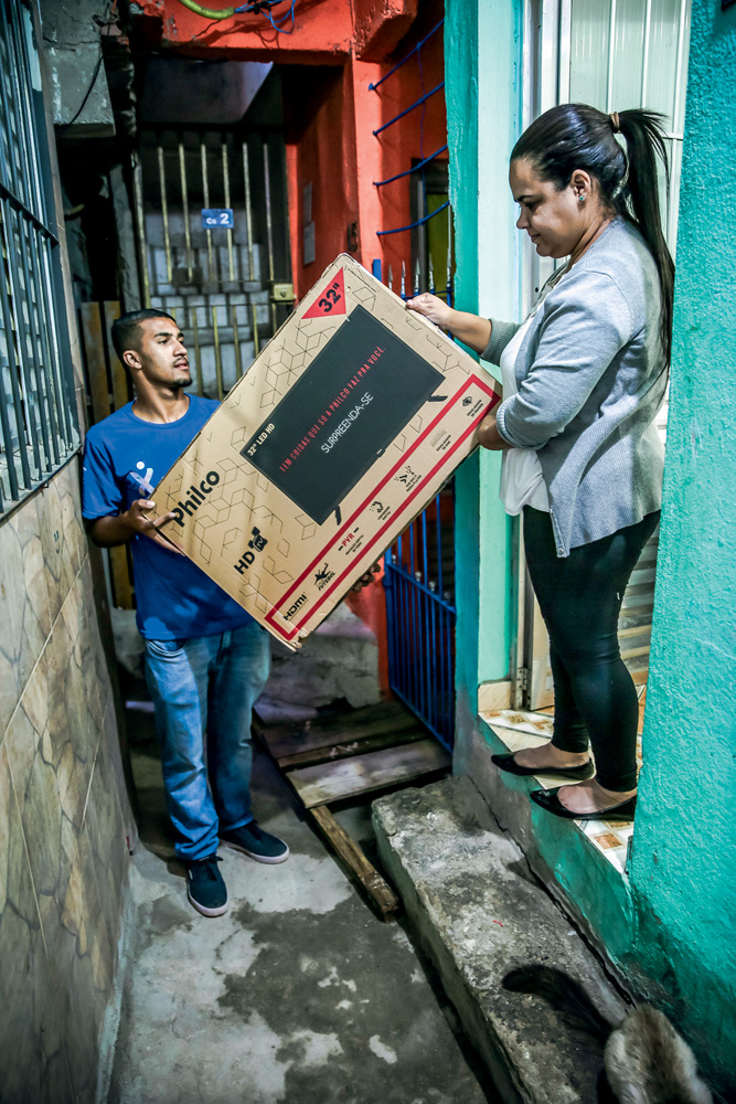 FAVELAEXPRESS_19.JPG Um entregador, com camiseta azul, seu uniforme, entrega uma grande caixa de papelão a um mulher na porta de uma casa, em Paraisópolis