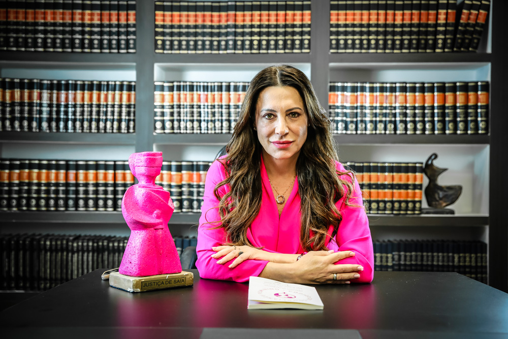 Gabriela Manssur veste camisa rosa e posa sentada de braços cruzados com livro em frente à mesa de madeira e uma boneca rosa ao lado. No fundo, livros de biblioteca.