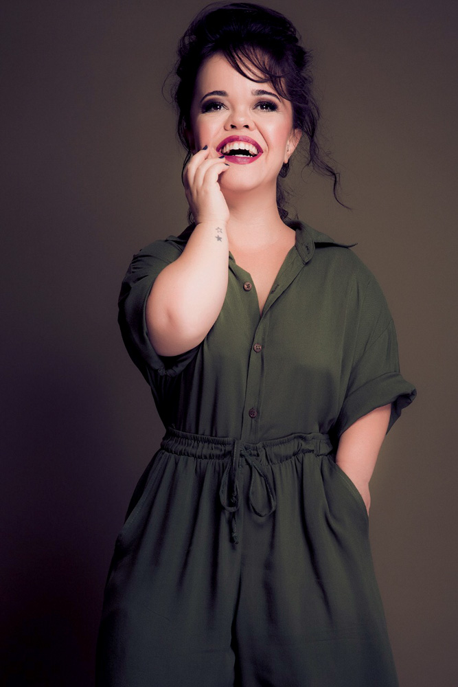 Juliana Caldas – Crédito Pino Gomes.JPG Juliana Caldas, mulher branca, magra e com nanismo, posa sorrindo, olhando para cima e com uma das mãos no queixo. Ela está em pé, tem cabelos presos e castanhos, e veste um macacão verde escuro