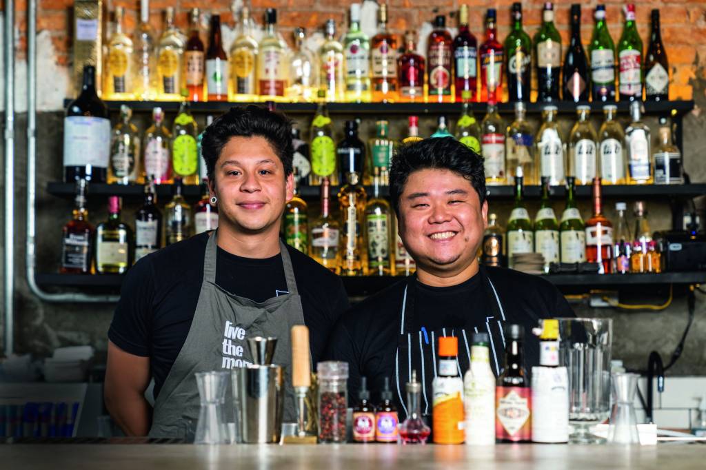Koya88_2020-11-25-65 – Kato.jpg Dois homens, um branco e outro com traços asiáticos, atrás de um balcão de um bar, com várias garrafas ao fundo