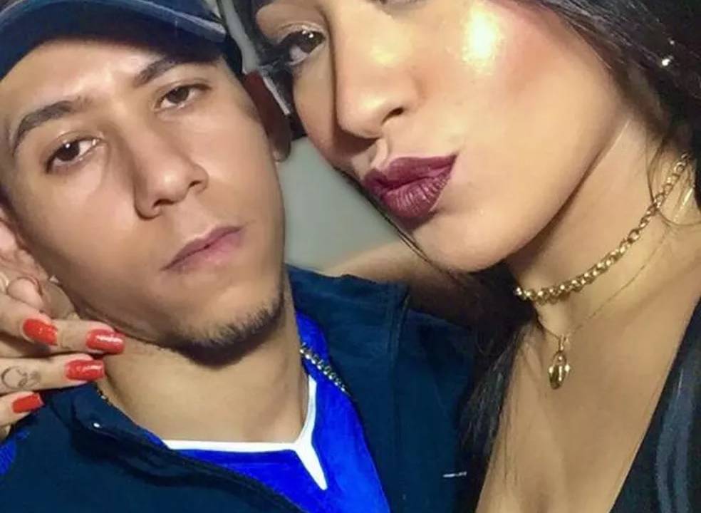 Foto exibe casal lado a lado. Ele à esquerda, com cara séria, camisa da seleção brasileira, boné e óculos espelhados em cima do boné. Ela de batom roxo e cabelo preto liso.