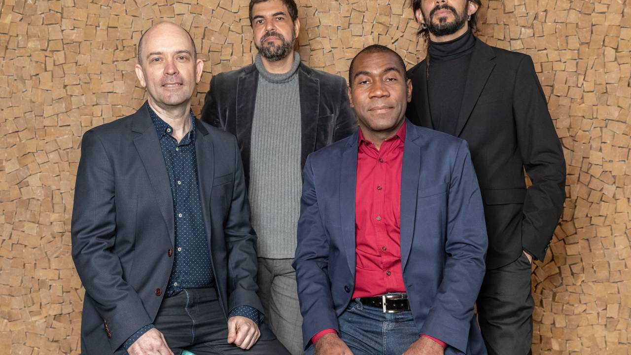 Quatro pianistas, todos homens, posam para uma foto em um fundo marrom. Ao fundo, em pé, dois homens brancos, de cabelos e barbas escuros, vestem roupa social preta e cinza. Na frente, sentados, dois homens carecas, um branco e outro negro, usando terno.